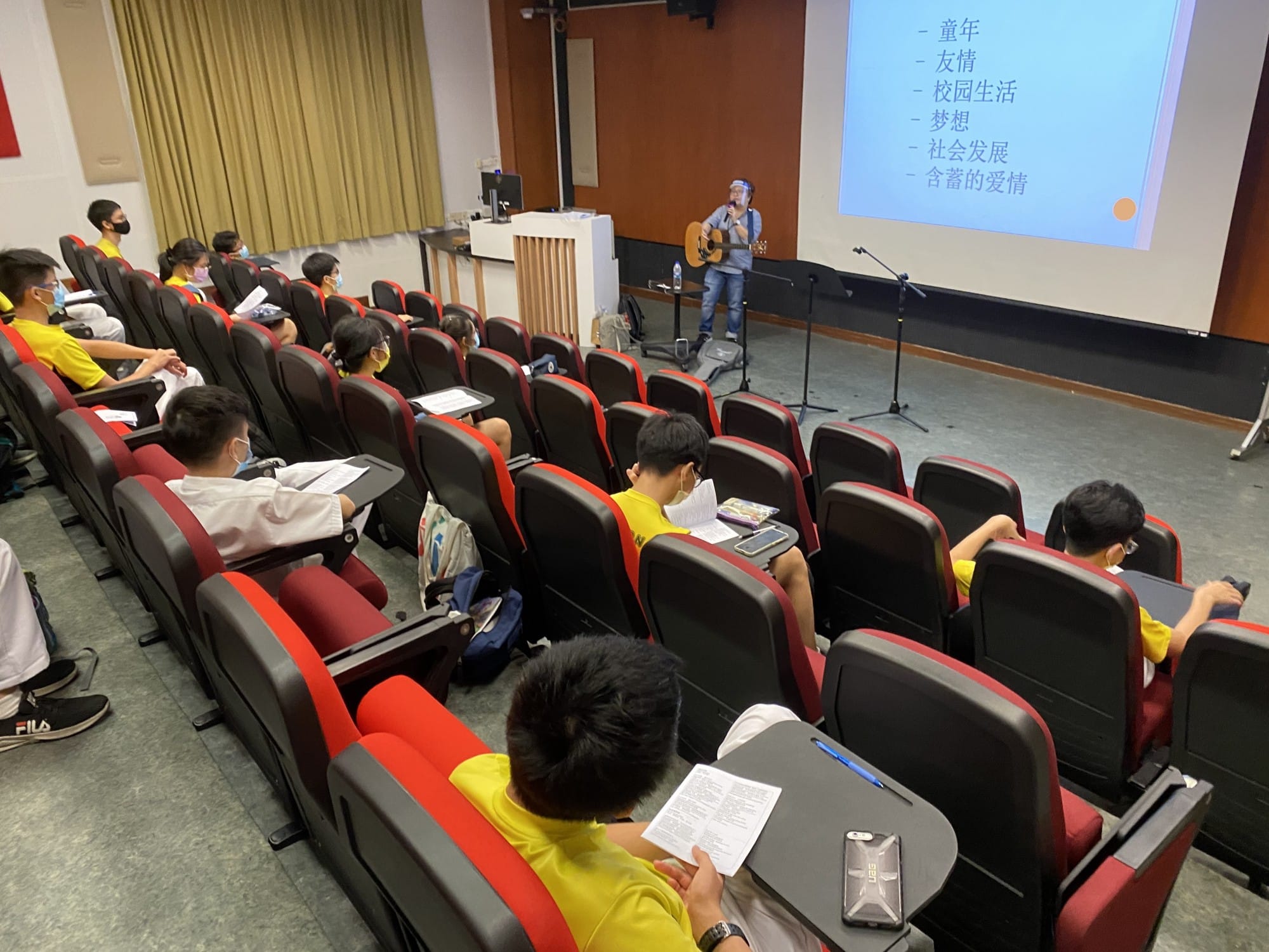 Students in red-black seats watch a guitarist presenting with a Chinese-text slideshow in a small lecture hall.